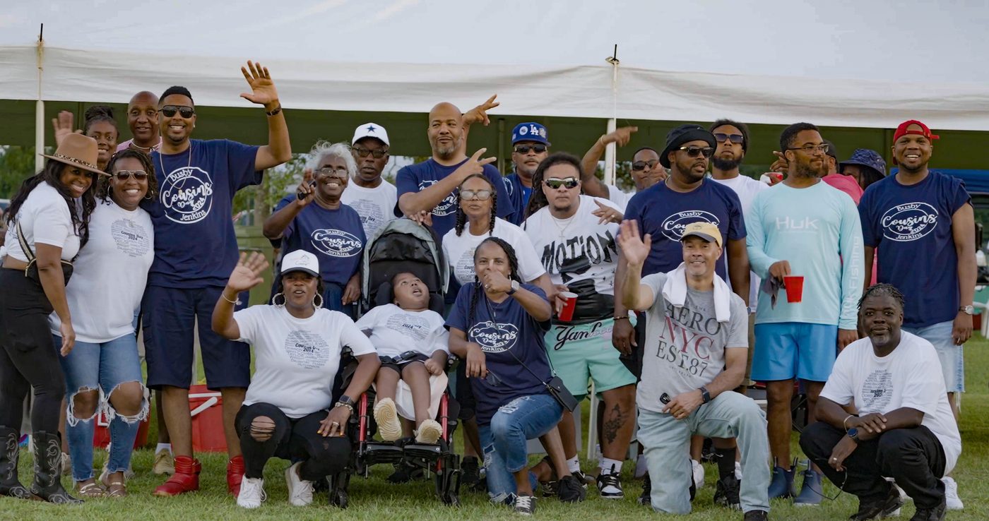 The Busby and Jenkins families waving in matching Cousins Day shirts under the tent